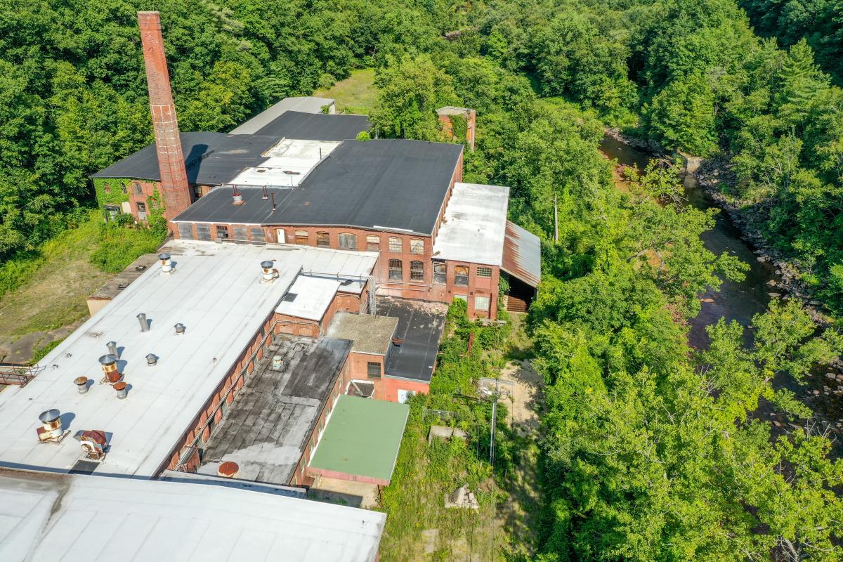 a drone photo of mismatched roofs of a mill complex on the left side, to the right is a stream and bright green forest