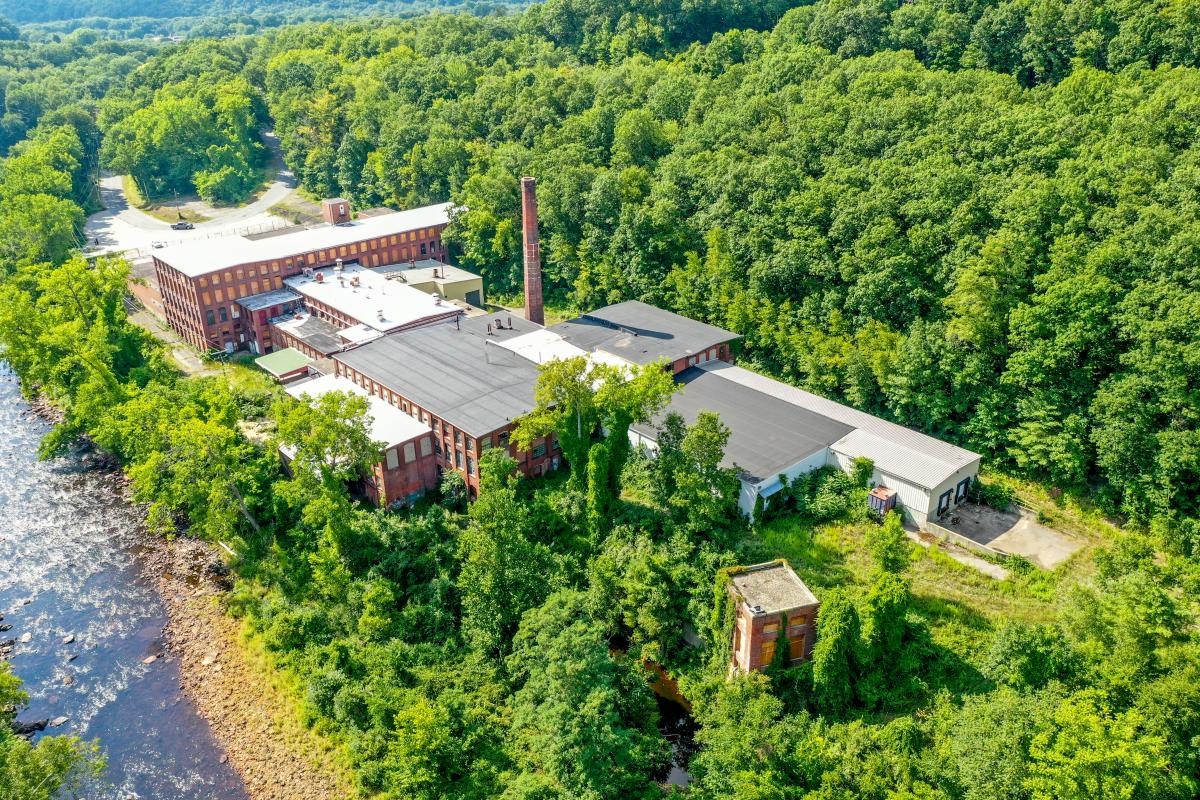 a mill building photographed by a drone, from the back of the complex, surrounded by forest and a stream