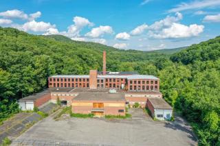 A drone photograph of the face of a large abandoned, brick mill building with green forest in the background