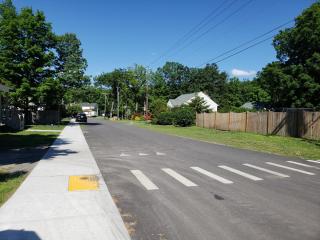A picture of River Street with a freshly paved road, a new sidewalk, and a crosswalk with a yellow ADA pad in the foreground