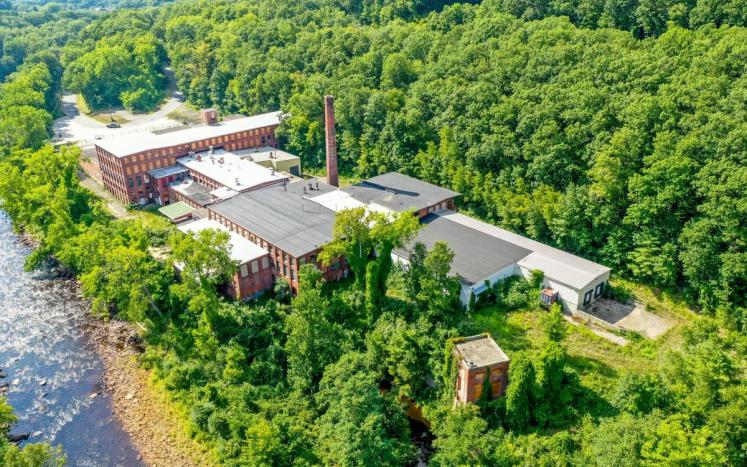 A panoramic drone view of an old mill complex surrounded by luch green trees and a beautiful river along the left side