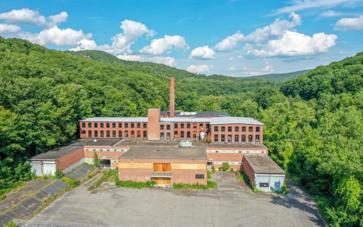 A drone photograph of the face of a large abandoned, brick mill building with green forest in the background